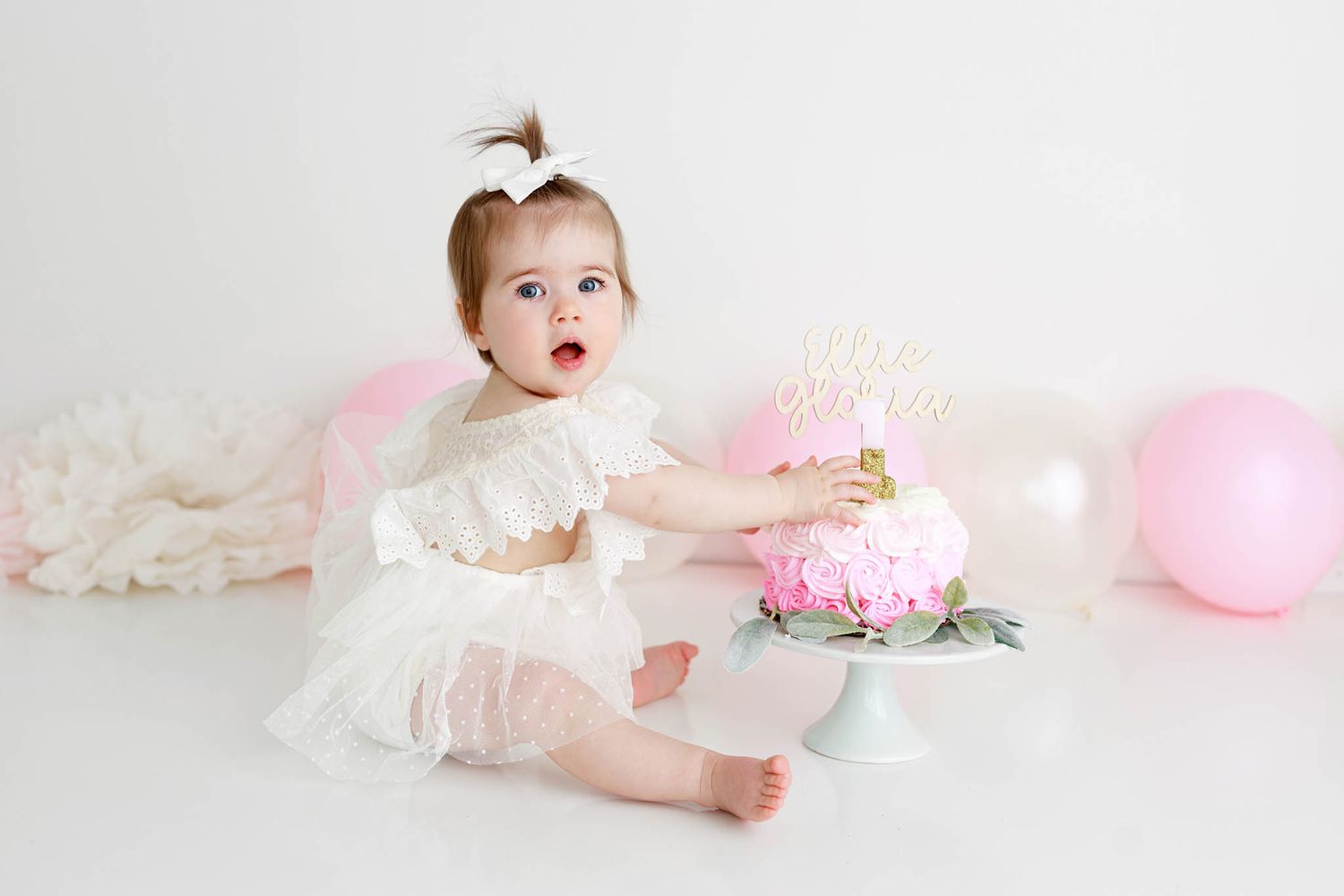 Menifee 1st birthday photographer: Baby girl in white dress reaching for a pink first birthday cake surrounded by balloons.
