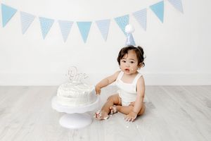 Winchester California 1st birthday photographer: Baby boy in white romper with first birthday cake beside blue bunting banner and party hat.