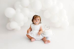 Baby girl in white dress sits with smash cake and white balloons during first birthday photo session.