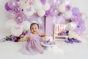 Baby girl in purple dress celebrates first birthday with lavender balloon arch, butterflies, and smash cake.