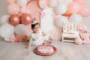 Baby girl in white dress sits near smash cake surrounded by pink roses and balloon arch for first birthday.