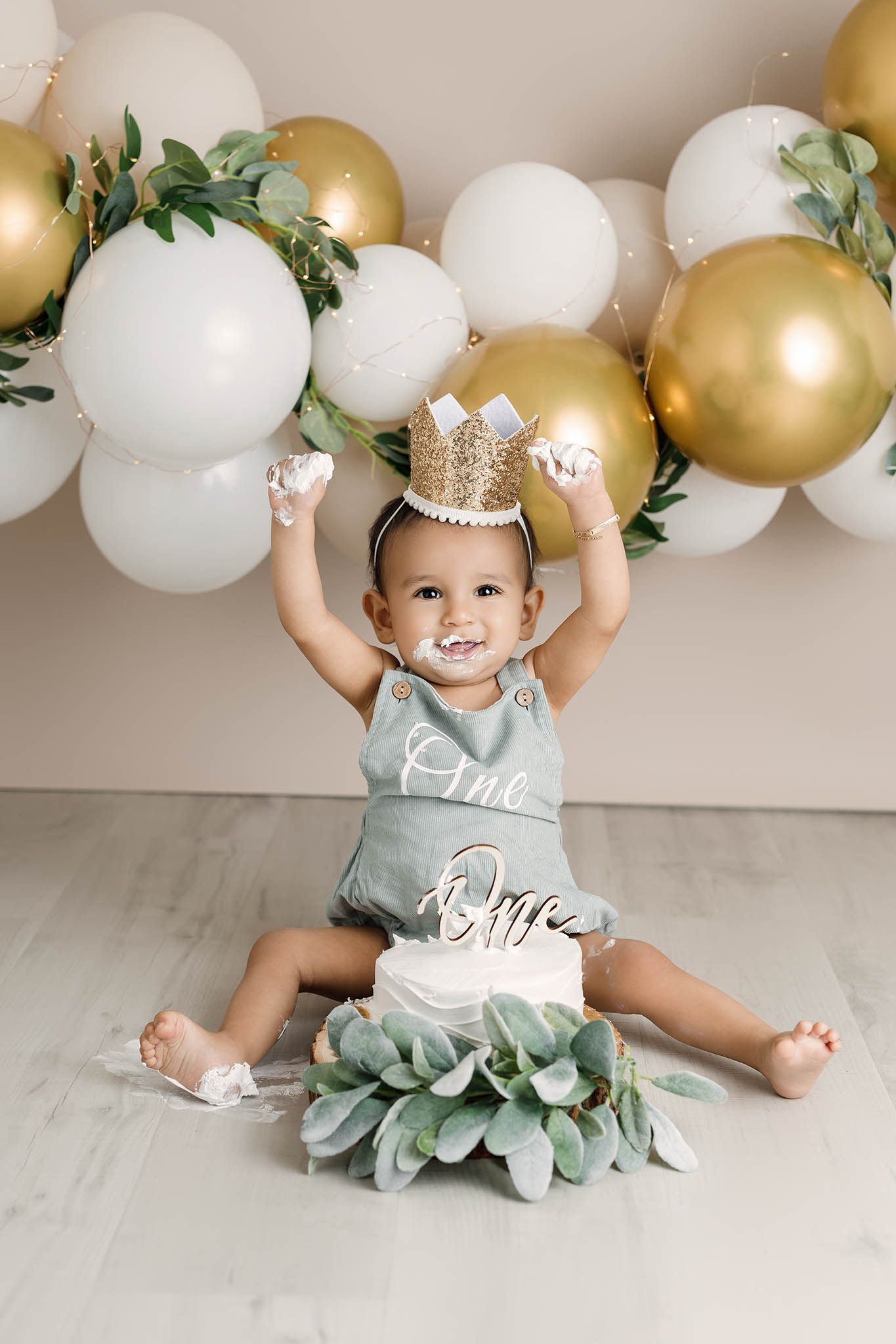 Happy baby in sage romper raises frosting-covered hands beside smash cake under gold and white balloon arch.