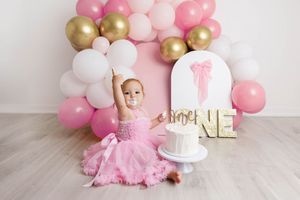 Baby girl in pink tutu dress reaches toward smash cake beside gold ONE letters and pink balloon arch.