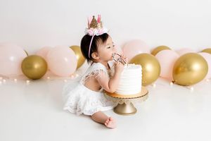Menifee cake smash photographer photo of baby girl in white dress and crown eating her first birthday cake surrounded by balloons.