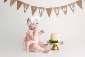 Baby girl in pink outfit with white bow sitting next to her first birthday cake under I'm ONE banner.