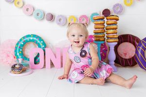 Happy baby girl in donut dress sitting among colorful donut decorations for first birthday cake smash.