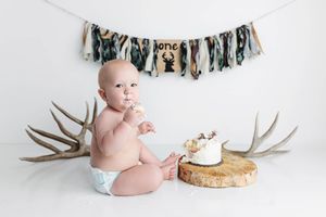 Baby boy in diaper sitting near first birthday cake on wood slice with antler decorations and one banner.
