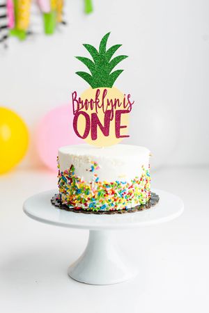 White birthday cake with colorful sprinkles and pineapple topper on cake stand.