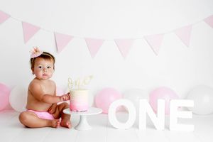 Baby girl in pink diaper with flower crown sitting next to pink cake beside large ONE letters and balloons.