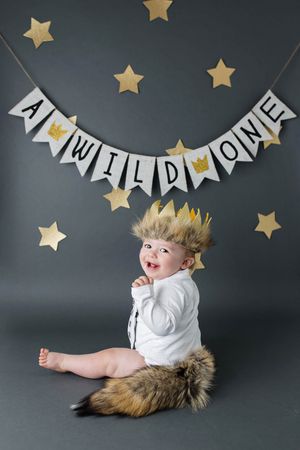 Baby in Wild One costume with fur crown and tail sits under A Wild One banner with gold stars.