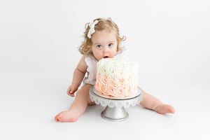 Curious baby girl bites into floral rosette cake on silver stand during first birthday cake smash session.