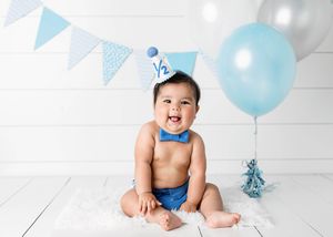 Smiling baby boy in blue bow tie and half birthday hat poses with balloon for 6 month photos.