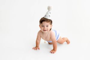 A smiling baby wearing a party hat and diaper crawls on a white background, celebrating their first birthday.