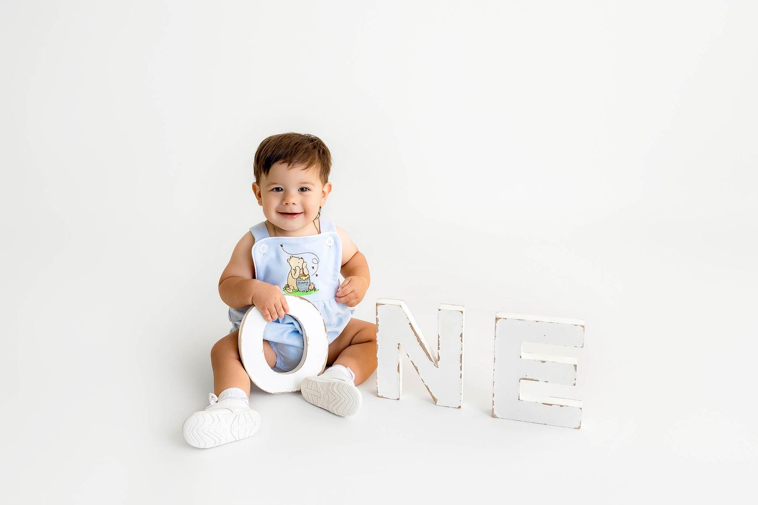 Smiling baby boy sitting with wooden ONE letters, wearing white outfit, celebrating his first birthday.
