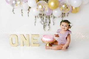 Baby girl in lavender dress sits beside pink smash cake with glittery ONE letters and balloon garland.