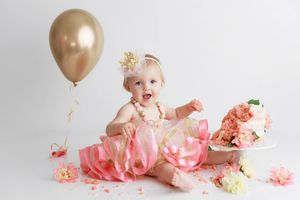 Adorable baby girl in pink tutu dress sits beside smashed floral cake with gold balloon for first birthday photo.
