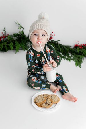 Baby in Christmas pajamas and knit hat drinking milk with cookies on white backdrop.
