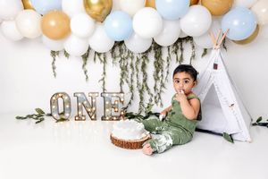 Baby boy in green outfit sits beside a cake at his ONE birthday cake smash with balloons and teepee.