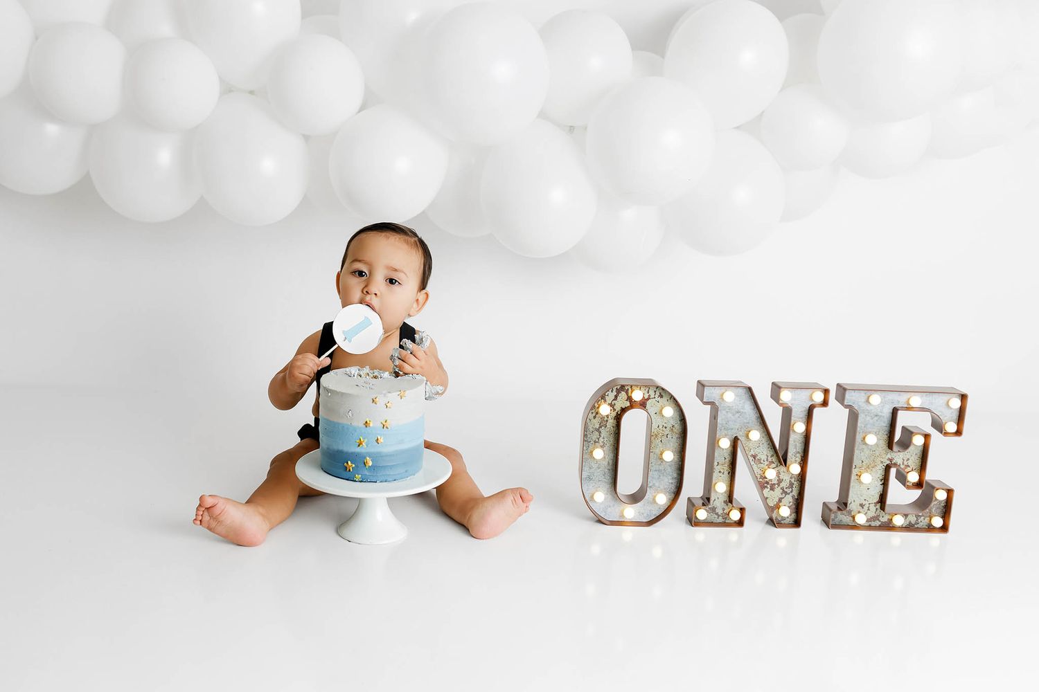 Murrieta 1st birthday photographer: Baby boy in diaper sitting with a blue ombre cake next to a ONE sign and white balloons.