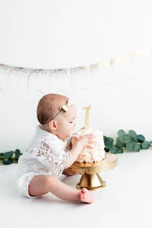 Baby in white lace outfit reaches into first birthday cake on gold stand with glitter number one topper.
