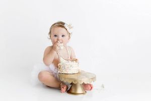 Baby girl in white tutu smashes first birthday cake on gold stand with white background.