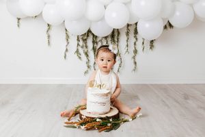 Perris 1st birthday photographer: Baby in white onesie sits with first birthday cake surrounded by florals and white balloon garland.