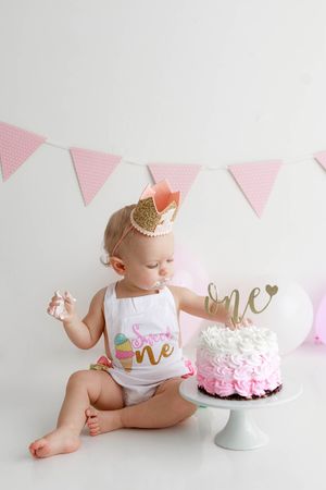 Baby girl in white romper with gold crown smashes pink ombre cake with one topper and pink bunting.