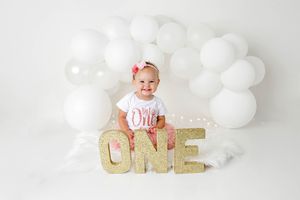 Wildomar 1st birthday photographer: Smiling baby girl in white ONE shirt sits before white balloon arch with gold glitter ONE letters.
