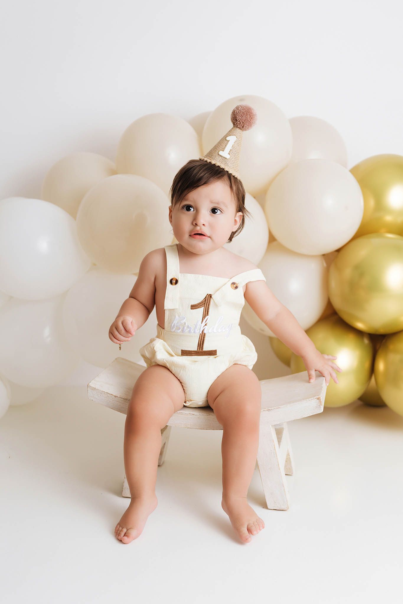 Baby in cream romper and party hat sitting on bench with white and gold balloon arch for first birthday.