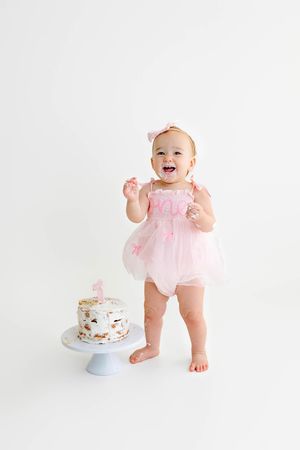 Baby girl in pink tutu romper smashing her first birthday cake on white background.