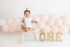 Baby girl in white dress sits in chair surrounded by pink balloons with glittery ONE letters for first birthday.