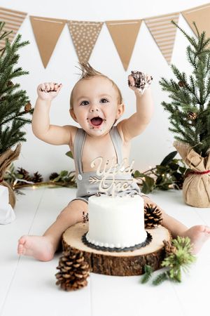 Excited baby boy celebrates first birthday smash cake session with Wild One cake and rustic pine tree decor.