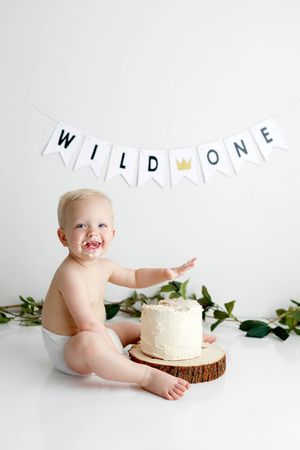 Happy baby in diaper smashes white cake on wood slice under Wild One banner with greenery decor.