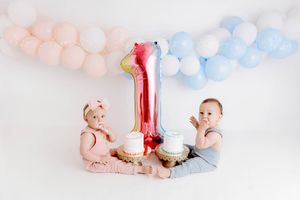 Twin babies celebrate their first birthday with smash cakes and a large number one balloon surrounded by pink and blue balloons.