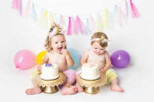 Twin baby girls in colorful tutus smash white cakes on gold stands with pastel balloons and bunting backdrop.