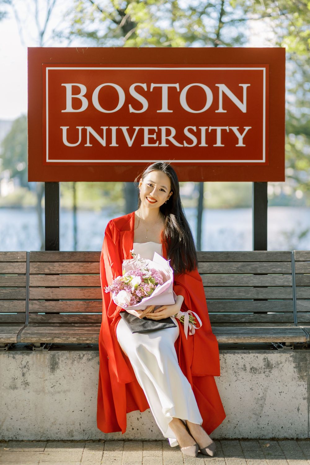 Boston University Graduate in red graduation gown holding pink flowers, sitting by Boston University sign on a sunny day.