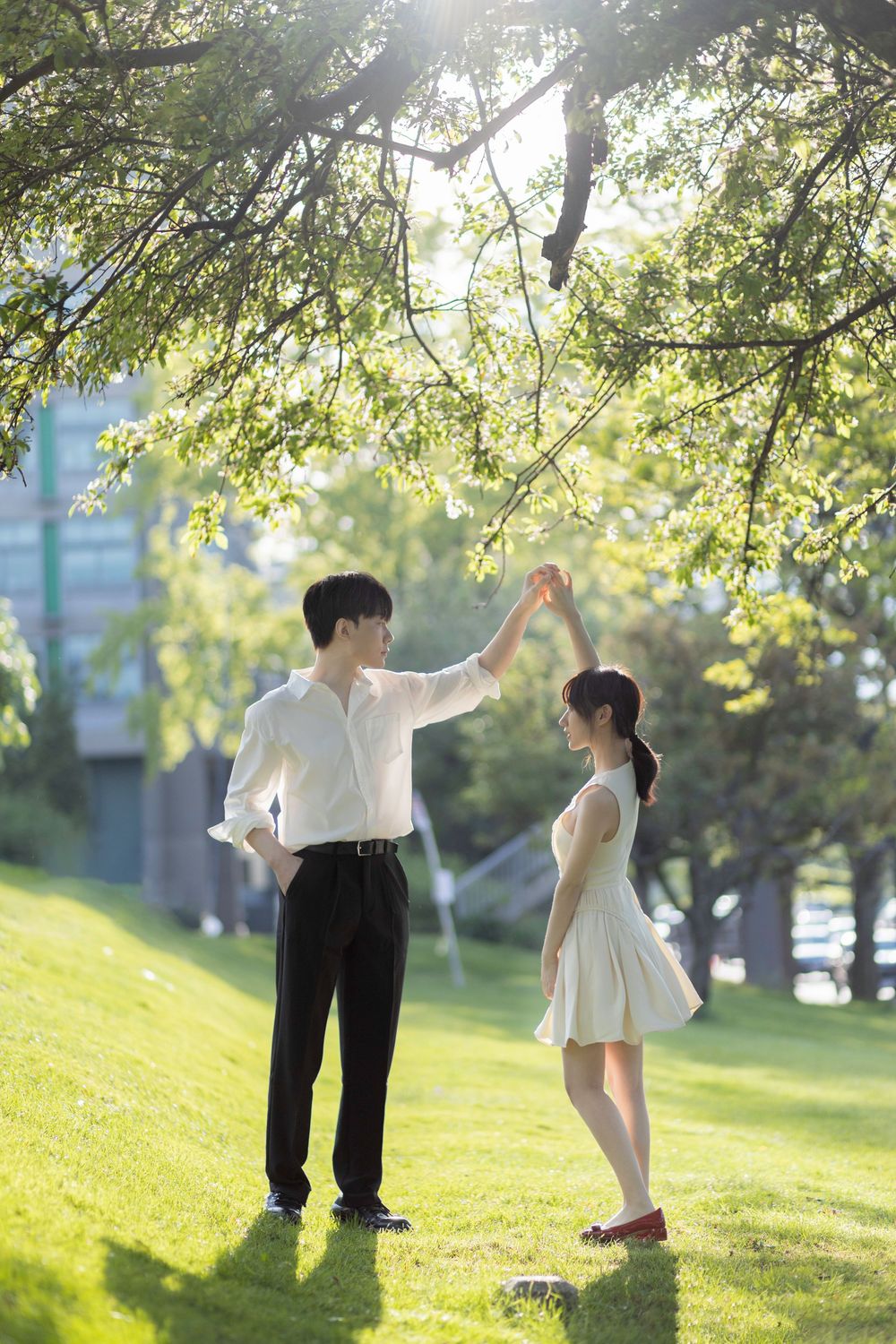 A young couple dancing together on green grass under a tree in a sunny park, dressed in white outfits.