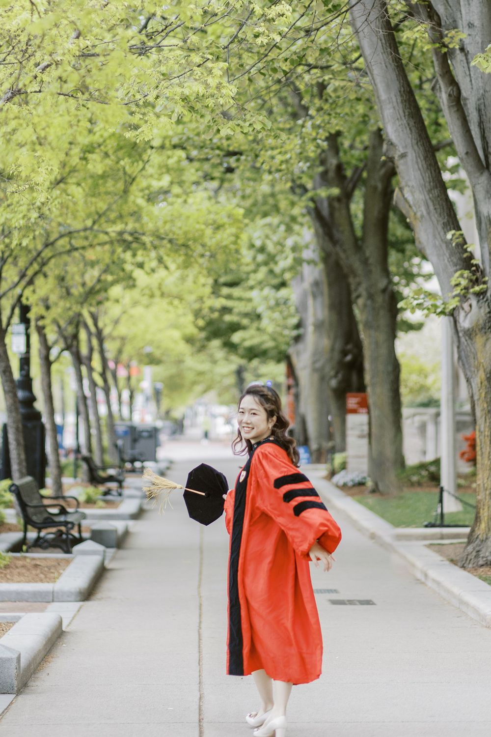 Boston University Graduate in red doctoral gown holds cap on tree-lined campus path during spring commencement celebration.