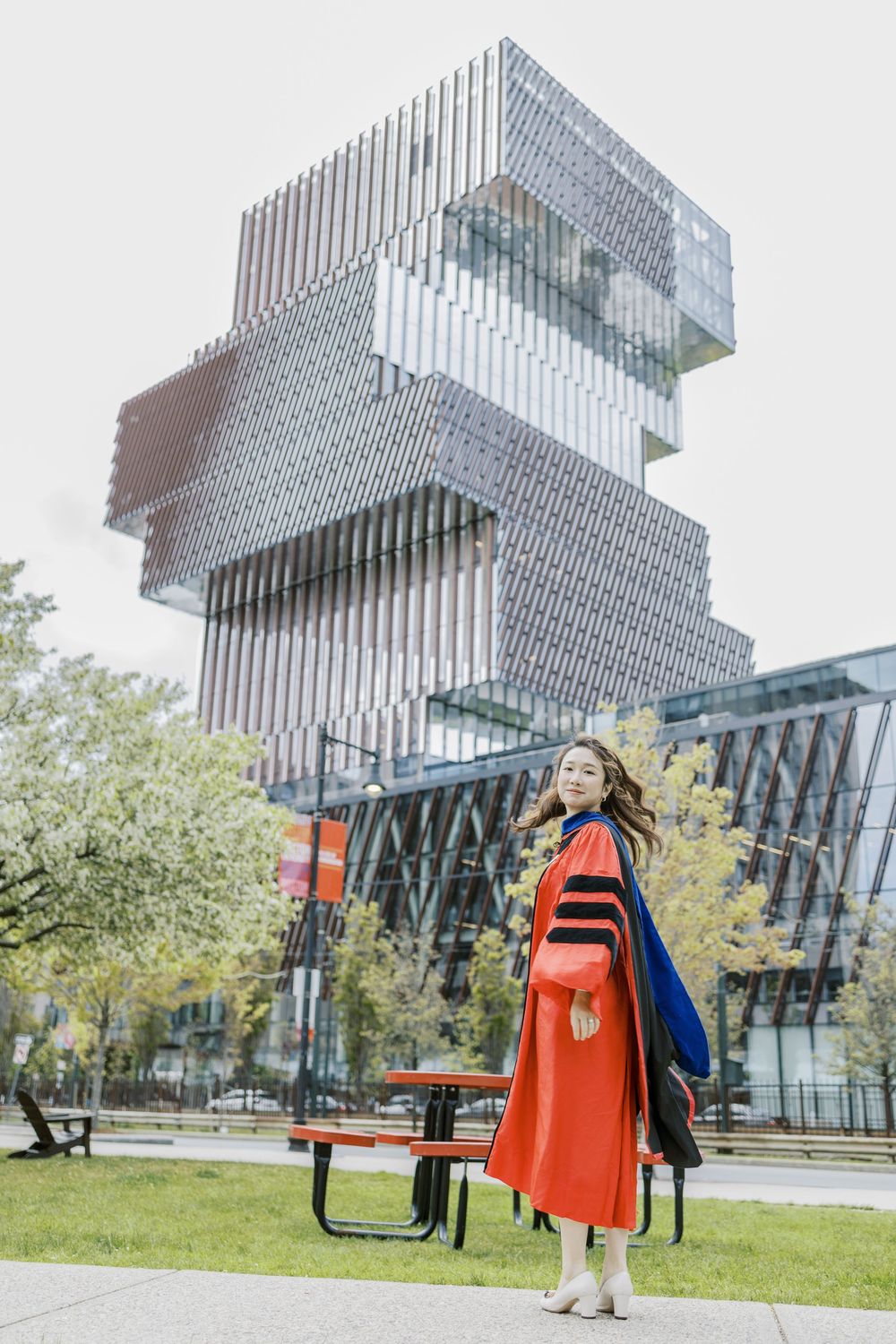 Boston University Graduate in red doctoral gown poses outdoors before a modern geometric glass building on a university campus.