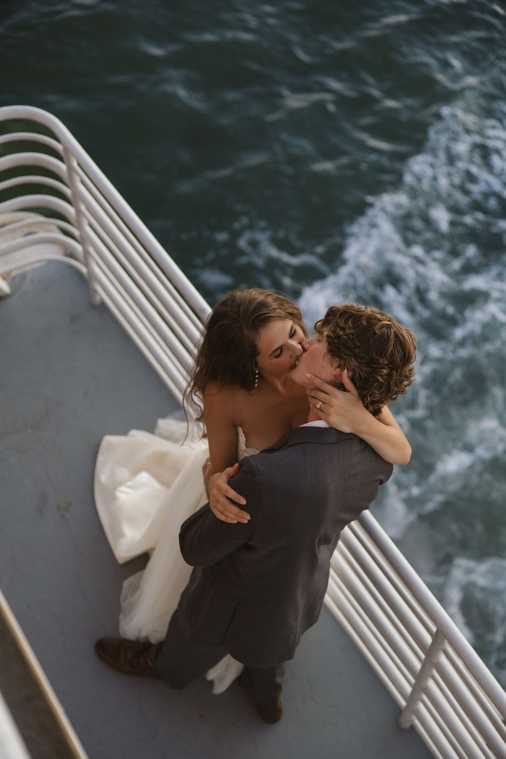 Bride in a white gown and groom in a gray suit kiss on a boat deck beside a railing, with ocean waves churning below.
