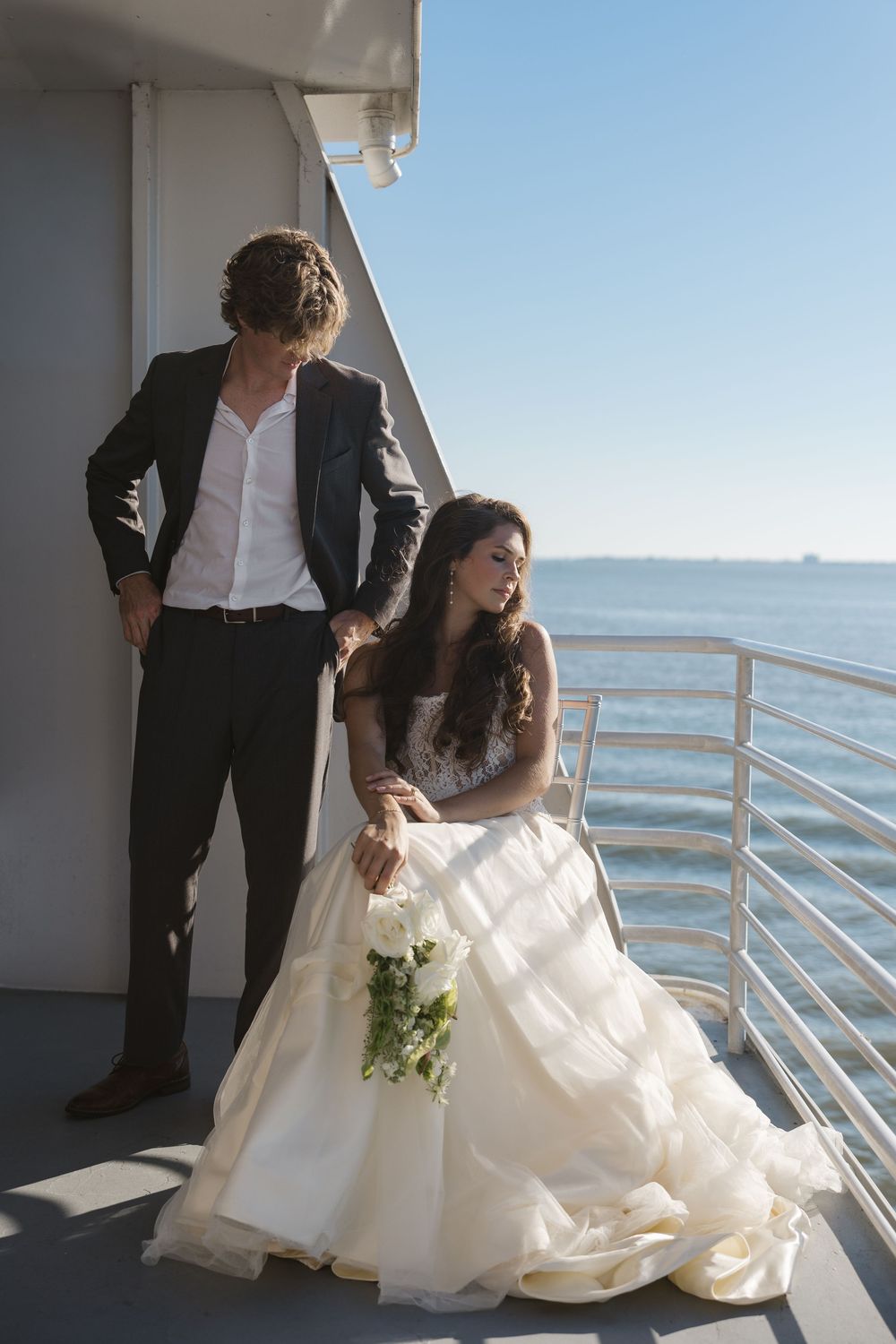 Bride in a flowing ivory gown holding a bouquet sits on a boat deck beside a standing groom in a dark blazer, with the sea behind.