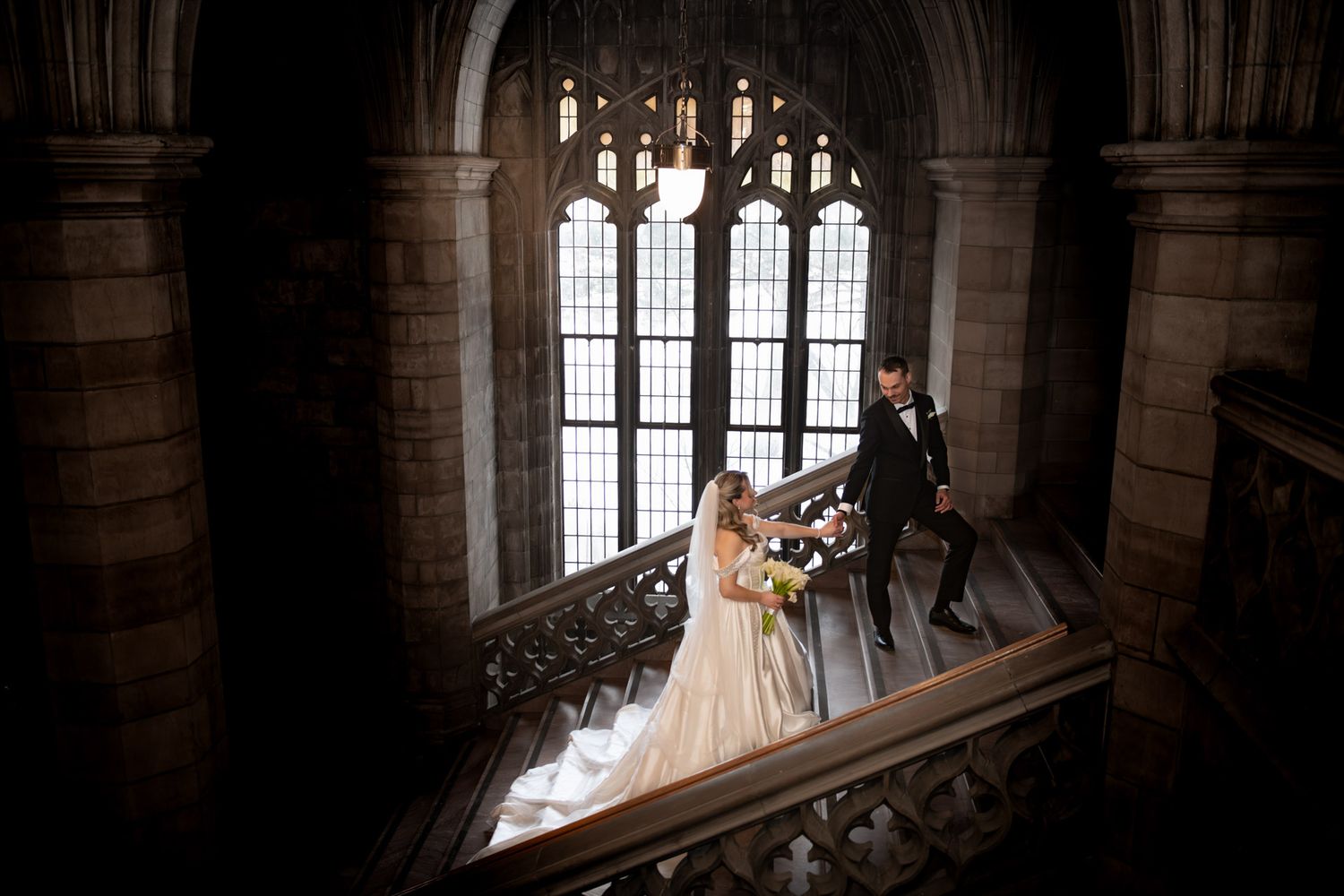 Bride and groom descend grand Gothic stone staircase with arched windows in dramatic wedding photo.