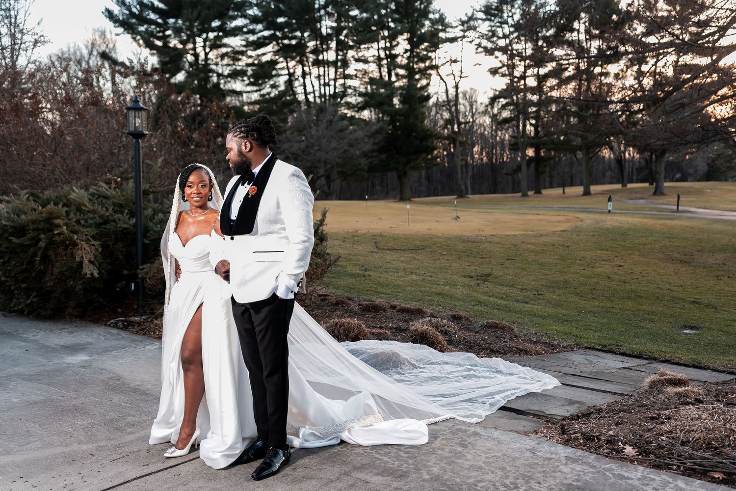 Elegant bride and groom in white wedding attire pose outdoors on a paved path with lush greenery backdrop. Deerfield Golf Club Newark De