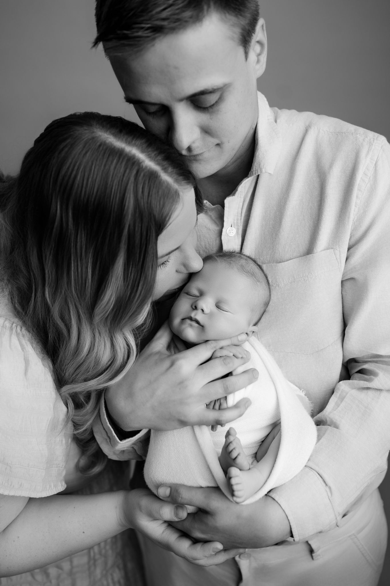 Black and white photo of parents tenderly holding and kissing their sleeping newborn baby in a studio portrait.