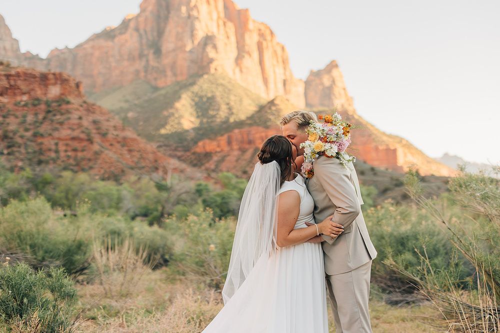 Bride and groom embracing with colorful bouquet before red rock cliffs at Zion National Park wedding.
