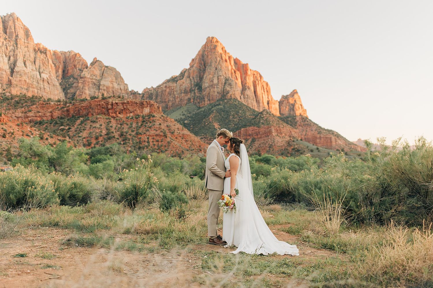 Bride and groom share a romantic kiss at Zion National Park with stunning red rock formations at golden hour.