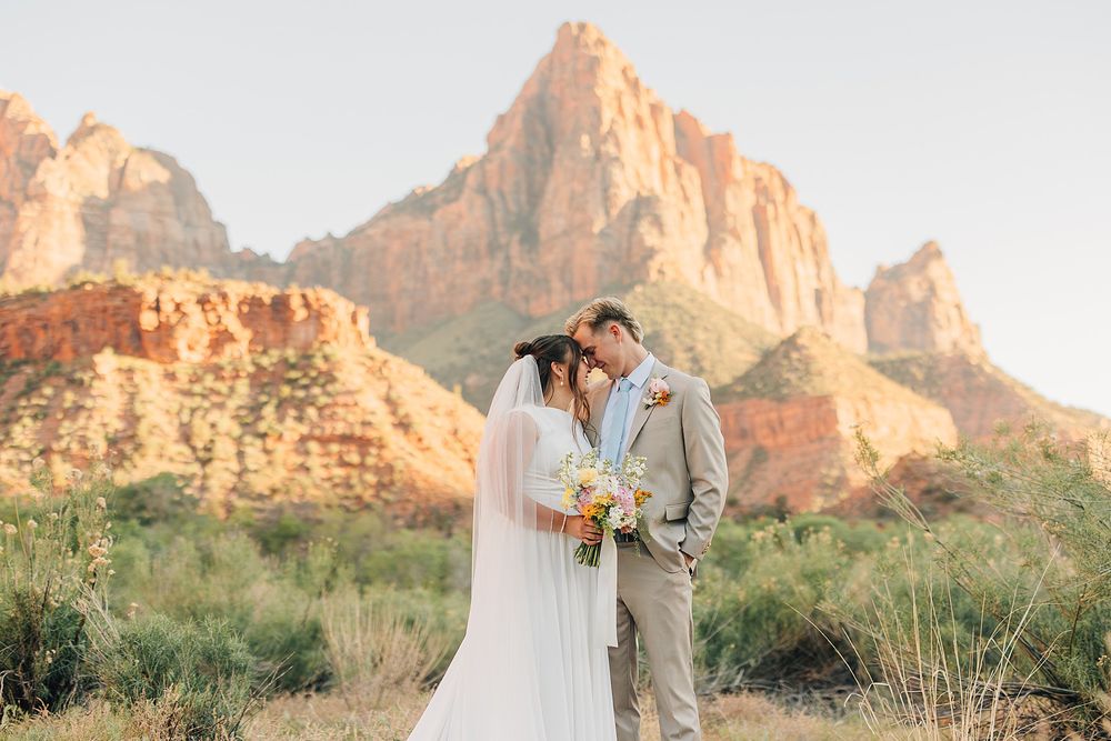 Newlyweds share intimate forehead touch with The Watchman mountain glowing at golden hour in Zion.