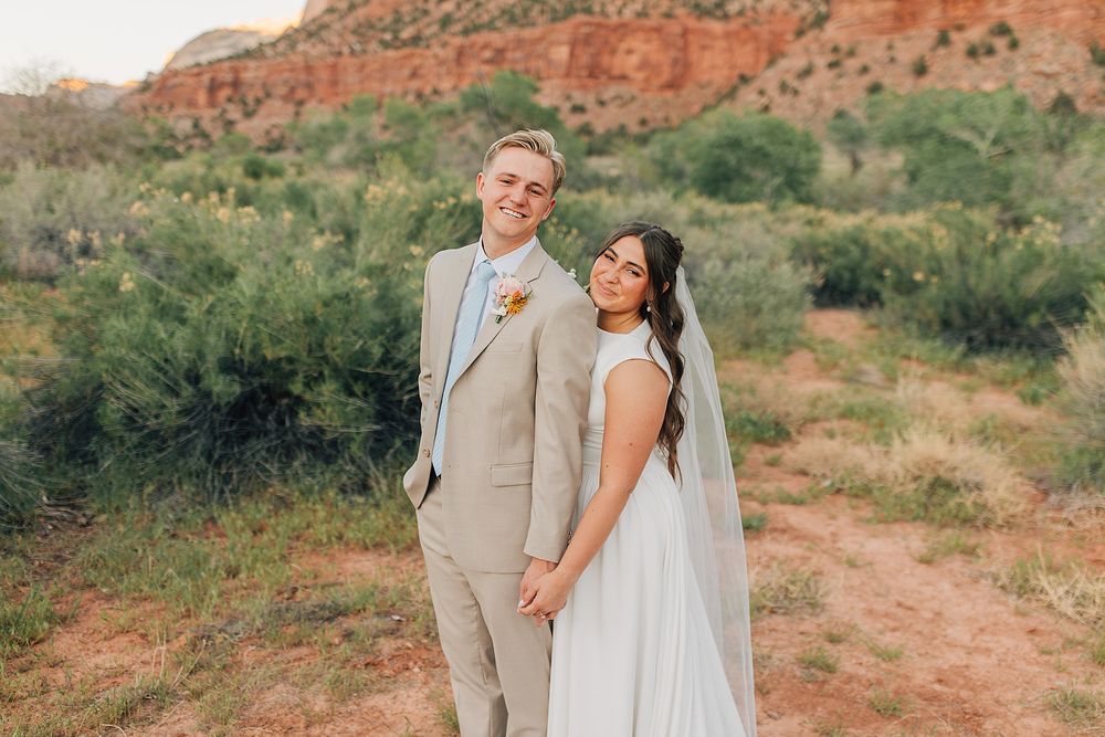 Bride and groom holding hands in front of red rock desert landscape, groom in tan suit, bride in white gown with veil.