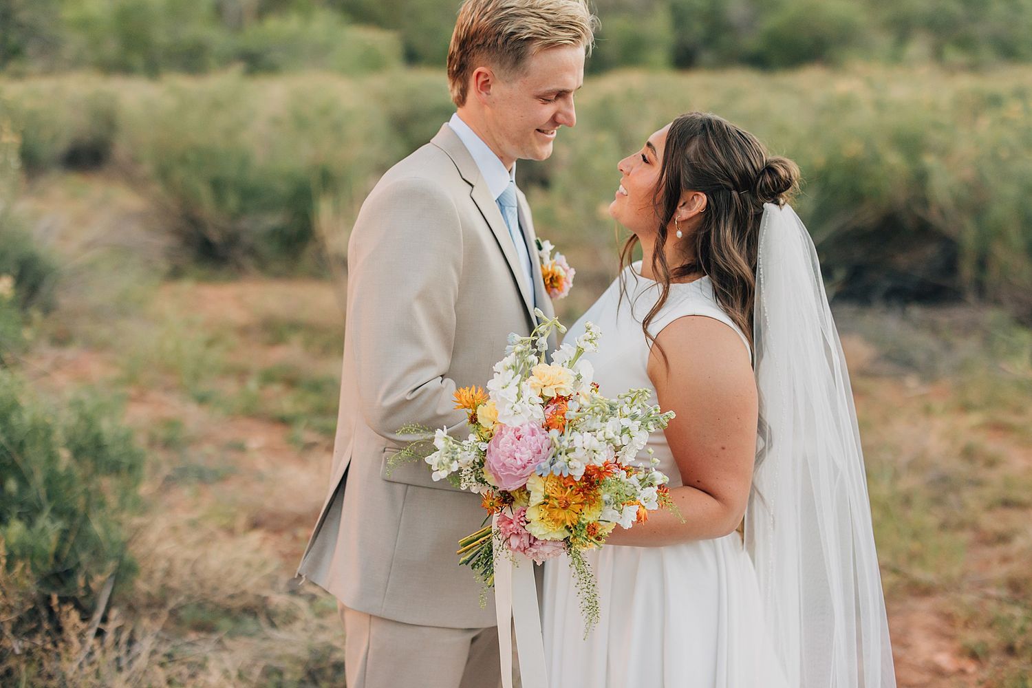 Bride and groom share a romantic gaze holding a vibrant wildflower bouquet in a desert landscape.