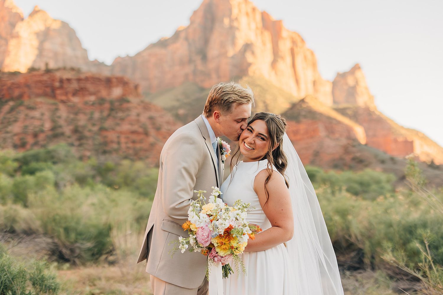 Groom kisses bride on cheek as she smiles holding bouquet at Zion National Park wedding.